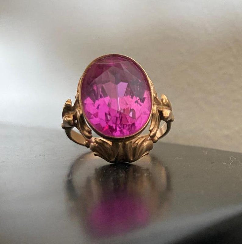 K&ouml;nnte beinhalten: Ein goldfarbener Ring mit einem gro&szlig;en, oval geschliffenen, facettierten rosa Edelstein. Der Ring hat ein verziertes, florales Design am Band und der Fassung. Der Edelstein reflektiert das Licht und erzeugt einen funkelnden Effekt.
