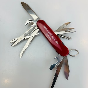 May include: A red multi-tool pocket knife with a variety of tools, including a saw, a bottle opener, a corkscrew, a screwdriver, and a pair of tweezers. The knife is open and the blade is extended. The knife has the text "Steinert, precisions, mbH Hamburg" printed on the handle.