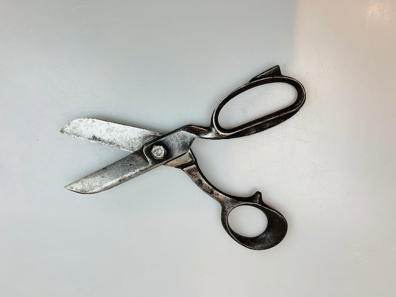 May include: A pair of heavy-duty metal shears with a dark, weathered patina on the handles and a central bolt. The blades are silver-toned with visible signs of wear, featuring a large oval finger loop and a smaller circular thumb loop.