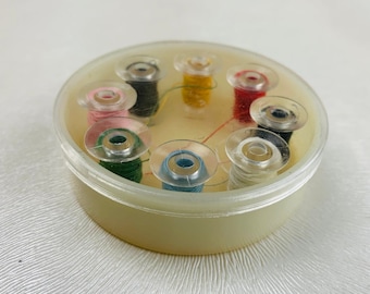 May include: A round, translucent plastic storage container holding eight individual sewing machine bobbins. Each bobbin is wound with thread in distinct colors, including pink, dark green, black, yellow, red, dark blue, light blue, and white.