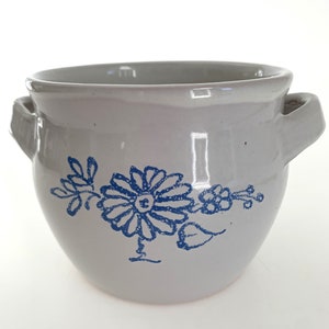 Puede incluir: Un tarro de cerámica blanco con dos asas y un diseño floral azul. El diseño presenta una flor grande con un pequeño grupo de flores y hojas.