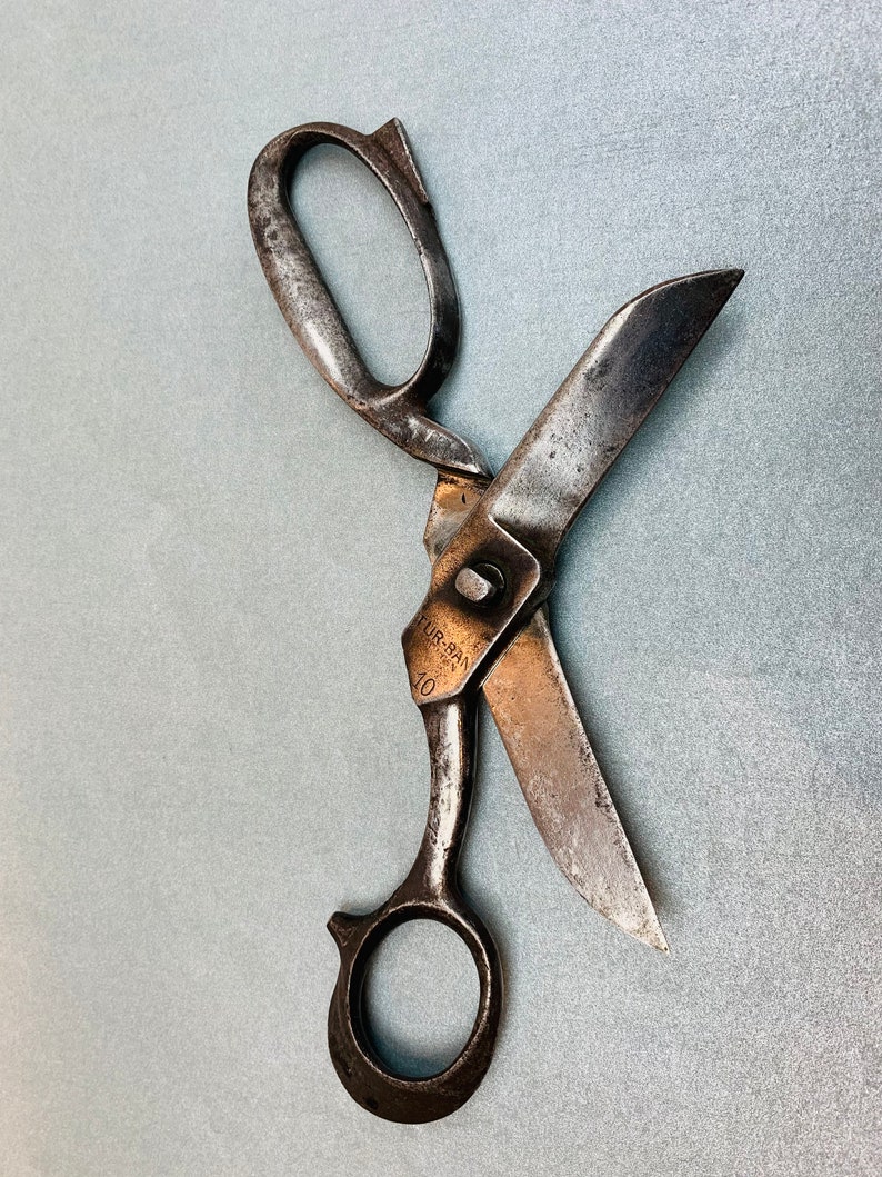 May include: A pair of vintage metal shears with a weathered, dark gray finish. The blades are partially open, and the pivot point is stamped with the text TUR-BAN and the number 10. The handles consist of one oval loop and one circular loop.