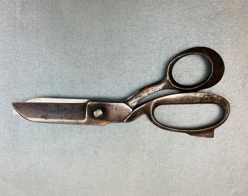 May include: A pair of vintage metal shears with a dark, weathered patina. The blades are straight and blunt-tipped, connected by a central screw. The handle features two oval finger loops, with the text "FUR-MAN" and the number "10" stamped on the metal.