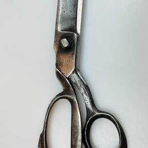 May include: A pair of heavy-duty metal industrial shears with a weathered, dark grey finish. The blades are straight and thick. The handle features two distinct loops, one larger than the other. The metal is stamped with the text TUR-MAN and 10.