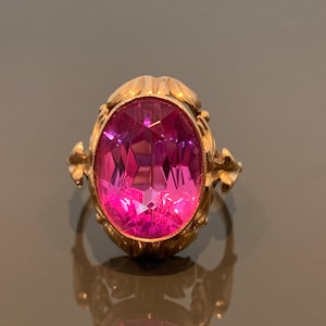 May include: A gold-toned ring featuring a large, faceted, oval-shaped pink gemstone. The ring has ornate detailing around the gemstone and on the band. The gemstone reflects light, creating a sparkling effect. Jewelry.