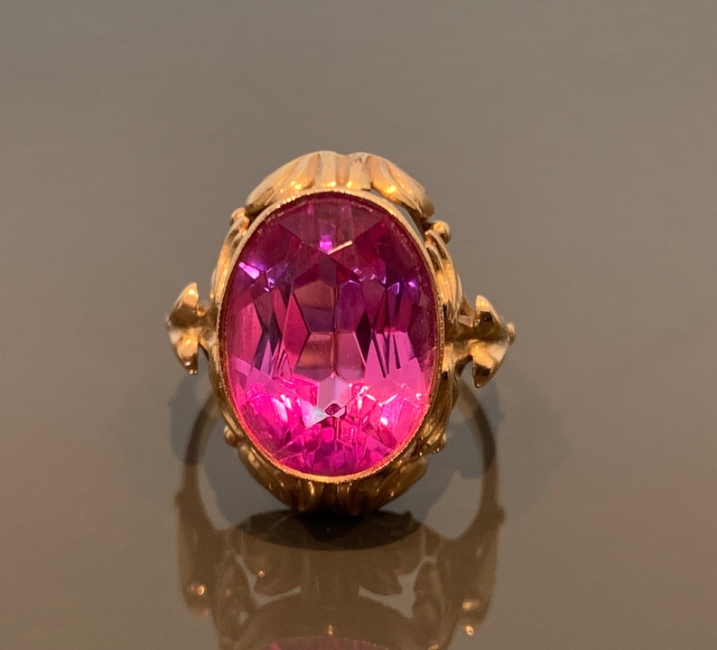K&ouml;nnte beinhalten: Ein goldfarbener Ring mit einem gro&szlig;en, facettierten, ovalf&ouml;rmigen, pinkfarbenen Edelstein. Der Ring hat verzierte Details um den Edelstein und am Band. Schmuck mit funkelndem Effekt.