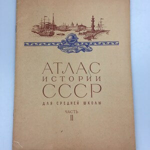 Puede incluir: Una cubierta de libro vintage con fondo marrón y texto azul y rojo. El título es "Atlas de la Historia de la URSS para la Escuela Secundaria Parte II". La cubierta presenta un dibujo de un horizonte de ciudad con barcos y un globo terráqueo.