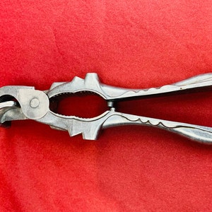 Può includere: Un schiaccianoci in metallo argentato con un design decorativo. Lo schiaccianoci ha una mascella incernierata e una leva per rompere le noci.