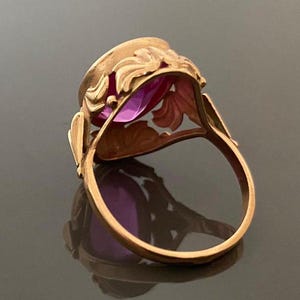 K&ouml;nnte beinhalten: Ein goldfarbener Ring mit einem gro&szlig;en, oval geschliffenen, lila Edelstein. Der Ring hat ein verziertes, florales Design um den Edelstein und an den Schultern des Rings. Der Ring ist vor einem dunklen Hintergrund platziert, der seine Details hervorhebt.