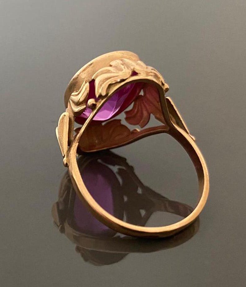 K&ouml;nnte beinhalten: Ein goldfarbener Ring mit einem gro&szlig;en, oval geschliffenen, lila Edelstein. Der Ring hat ein verziertes, florales Design um den Edelstein und an den Schultern des Rings. Der Ring ist vor einem dunklen Hintergrund platziert, der seine Details hervorhebt.