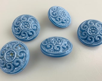 May include: Five round, light blue buttons featuring a raised, intricate swirl and dot pattern on the surface. Each button has a smooth, circular edge and a glossy finish, displaying a detailed relief design that radiates from the center.