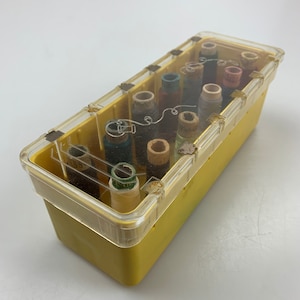 Puede incluir: Una caja de plástico amarilla con una tapa transparente que contiene carretes de hilo de varios colores. La caja tiene un patrón de cuadrícula y agujeros para ventilación.