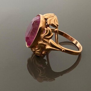 K&ouml;nnte beinhalten: Ein goldfarbener Ring mit einem ovalen, facettierten, rosafarbenen Edelstein. Der Ring hat ein verziertes, florales Design an den Seiten und um den Edelstein herum. Der Ring ist vor einem dunklen Hintergrund platziert, wobei sich sein Spiegelbild zeigt.