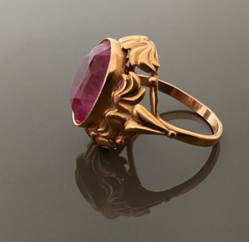 K&ouml;nnte beinhalten: Ein goldfarbener Ring mit einem ovalen, facettierten, rosafarbenen Edelstein. Der Ring hat ein verziertes, florales Design an den Seiten und um den Edelstein herum. Der Ring ist vor einem dunklen Hintergrund platziert, wobei sich sein Spiegelbild zeigt.