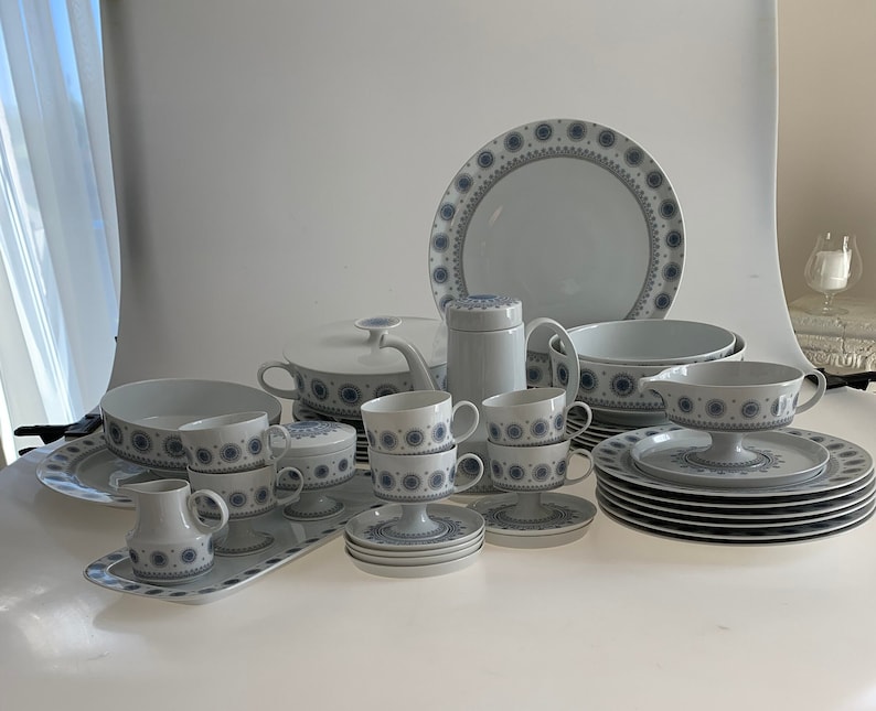 K&ouml;nnte beinhalten: Ein Set aus wei&szlig;em Porzellan-Geschirr mit einem blauen und grauen Blumenmuster. Das Set enth&auml;lt Teller, Schalen, Tassen, Untertassen, eine Teekanne, eine Zuckerdose, eine Milchk&auml;nnchen und eine Servierplatte.
