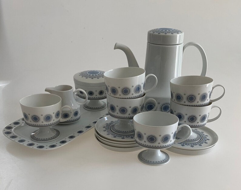 K&ouml;nnte beinhalten: Ein wei&szlig;es Teeservice mit blauen und grauen Blumenmustern. Das Set beinhaltet eine Teekanne, eine Milchk&auml;nnchen, eine Zuckerdose und vier Tassen mit Untertassen.