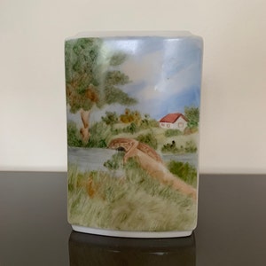 May include: A white ceramic vase with a hand-painted scene of a small house with a red roof, a bridge over a river, and green trees. The vase has a rounded rectangular shape.