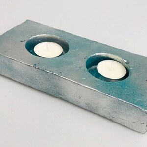 Puede incluir: Un portavelas de hormigón de color turquesa y plata con dos recortes circulares para velas votivas.