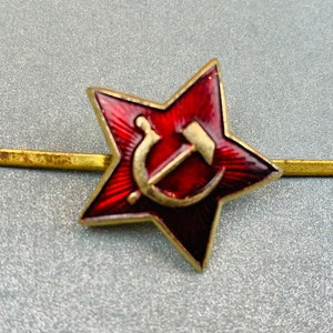 May include: A five-pointed star badge featuring a deep red enamel finish with a sunburst pattern. Centered on the star is a gold-toned hammer and sickle emblem. The badge is attached to a horizontal gold-toned metal bar that extends from both sides.