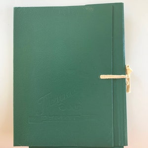 May include: A green folder with the text "Tanma" and "BY MAF" embossed on the front. The folder is tied with a beige ribbon.