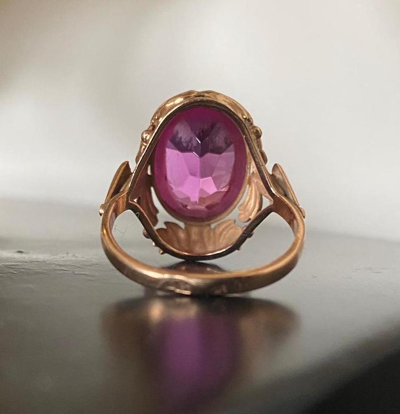 K&ouml;nnte beinhalten: Ein Vintage-Goldring mit einem ovalen, facettierten rosa Edelstein. Der Ring hat ein verziertes Filigran-Design um den Edelstein und auf den Schultern des Bandes. Der Ring ist auf einer dunklen Oberfl&auml;che platziert.