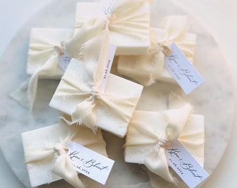 May include: White favor boxes tied with white ribbon and a small white tag with the text "Kiran & Blair" and the date "21.06.2024".