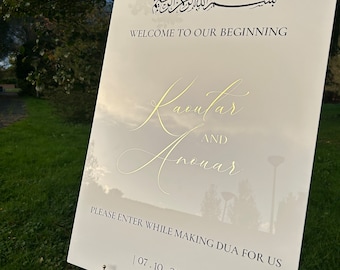Può includere: Un cartello bianco con lettere dorate che annunciano un matrimonio. Il cartello recita "Benvenuti al nostro inizio" con i nomi "Kaoular e Anouar" in calligrafia elegante. La data "07.10.2023" è anche inclusa. Il cartello è su un cavalletto dorato.