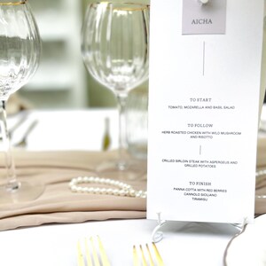 Op de afbeelding: Een witte menukaart met een gouden standaard, met een viergangenmenu voor een restaurant genaamd "AICHA". Het menu bevat "Om te beginnen", "Om te volgen", "Gegrilde biefstuk" en "Om te eindigen".
