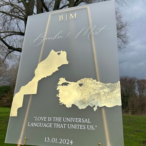 May include: A frosted acrylic wedding sign featuring gold foil maps of Morocco and Turkey. The sign displays the names "Benchra & Mert", initials "BM", the date "13.01.2024", and the quote "LOVE IS THE UNIVERSAL LANGUAGE THAT UNITES US."