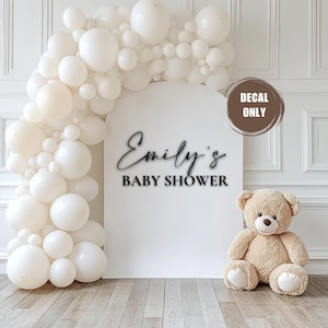 May include: White balloon arch with a white backdrop and black text that reads "Emily's Baby Shower".