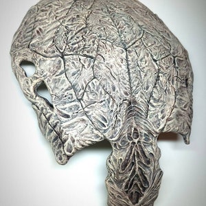 Könnte beinhalten: Eine weiße und graue, strukturierte Maske mit einem detaillierten Design. Die Maske hat eine spitze Unterseite und eine große Öffnung oben.