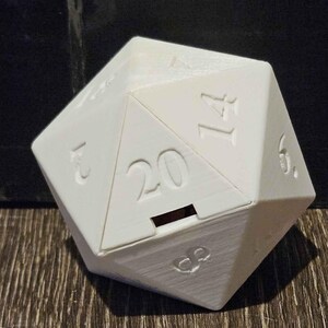 May include: A white 20-sided die with a clear plastic lid and a red slot. The die is sitting on a wooden surface next to a red and orange 20-sided die with black numbers.