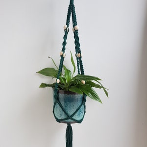 May include: A dark green macrame plant hanger with wooden beads and a tassel. A green potted plant is hanging in the macrame.
