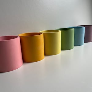 May include: Six cylindrical planters in a rainbow colour scheme, from pink to purple. The planters are made of a smooth, matt material and are arranged in a row from left to right.
