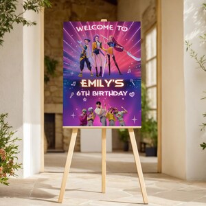 May include: A wooden easel displays a vibrant birthday sign. The sign reads "WELCOME TO EMILY'S 6TH BIRTHDAY" with colorful graphics of animated characters. The background is a gradient of purple and blue with star accents.