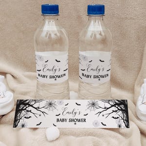 May include: Two clear water bottles with blue caps and labels that read "Emily's Baby Shower." The bottles are next to a banner with the same text and Halloween-themed graphics. White baby booties with bunny faces are on either side.