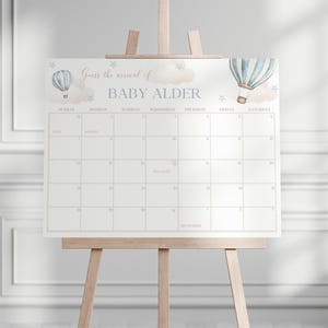 May include: A light-colored calendar on a wooden easel, featuring a design with hot air balloons and the text "Guess the arrival of BABY ALDER." The calendar grid is for tracking the baby's due date.