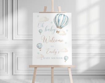 May include: A white rectangular sign featuring watercolour illustrations of blue and white hot air balloons, clouds, and stars. The text reads Oh baby Welcome TO Emily's BABY SHOWER in a mix of blue and brown script and block lettering fonts.