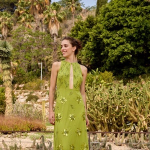 May include: A woman wearing a green halter-neck maxi dress with floral embroidery. The dress has a keyhole opening at the neckline and is made of a sheer fabric. The woman is standing in a garden setting with green plants and trees in the background.