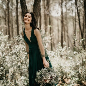 Könnte beinhalten: Eine Frau in einer Waldkulisse, die ein dunkelgrünes Kleid und braune Stiefel trägt. Sie hält einen Strauß weißer und grüner Blumen.