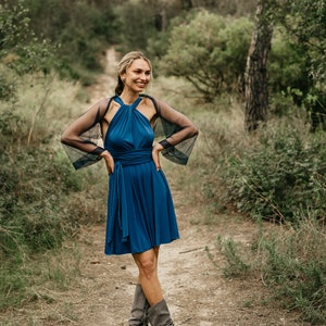Könnte beinhalten: Eine Frau in einem blauen Wickelkleid mit einem schwarzen, durchsichtigen Überwurf und braunen Stiefeln. Sie steht auf einem Feldweg in einem Wald.