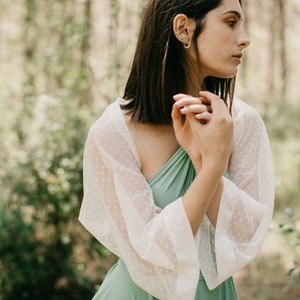 Könnte beinhalten: Eine Frau trägt ein salbeigrünes Kleid mit einer weißen, gepunkteten, durchsichtigen Überlage. Das Kleid hat einen einseitigen Schnitt und die Überlage hat lange Ärmel.