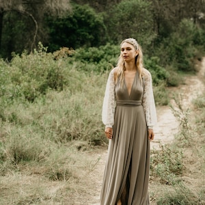 Könnte beinhalten: Eine Frau in einem langen, fließenden, taupefarbenen Kleid mit einem weißen Spitzenüberzug an den Ärmeln. Sie steht in einer Waldlandschaft mit grünem Laub und einem Erdweg.