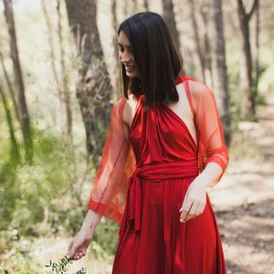 Könnte beinhalten: Eine Frau in einem roten Kleid mit einem durchsichtigen roten Überwurf steht in einem Wald. Das Kleid hat einen Halterneck und eine Schleife in der Taille.