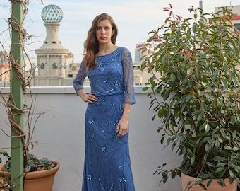 May include: A woman wearing a long, blue, beaded dress with a scoop neckline and three-quarter sleeves. She is standing on a wooden deck with potted plants in the background.