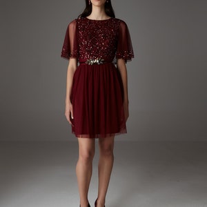 Könnte beinhalten: Ein burgunderfarbenes Tüllkleid mit einem Paillettenoberteil und einem goldenen Blattgürtel. Das Kleid hat kurze Ärmel und einen ausgestellten Rock.