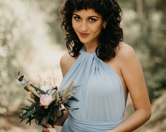 May include: A woman in a light blue halter dress holds a bouquet of flowers with a white lace ribbon. She is standing in a forest setting.