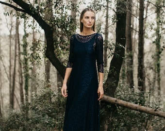 May include: A woman in a dark blue lace dress stands in a forest. She is looking to the right of the frame. The dress has a scoop neckline and three-quarter sleeves.