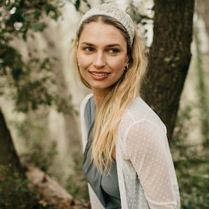 Könnte beinhalten: Eine Frau mit langen blonden Haaren trägt einen weißen Cardigan mit weißen Punkten und ein silbernes Stirnband. Sie steht in einem Wald.