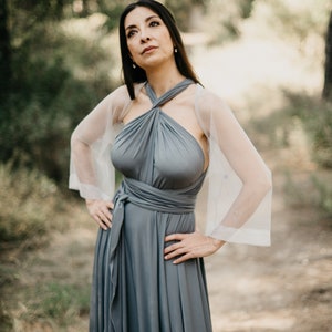 Könnte beinhalten: Eine Frau trägt ein graues Maxikleid mit einem weißen, durchsichtigen Schal. Das Kleid hat einen Halterneck und ist in der Taille gebunden. Die Frau steht in einer Waldlandschaft.